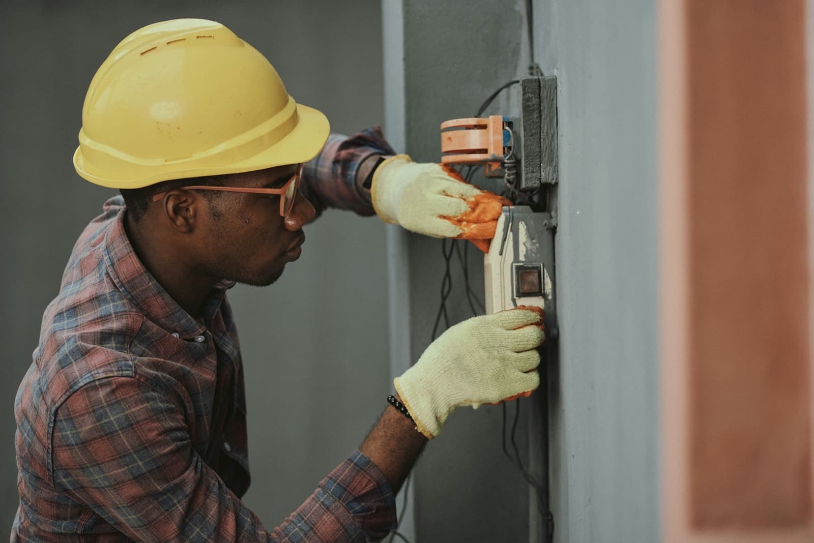 An electrician in a yellow hard hat and gloves testing a wall mounted electrical box