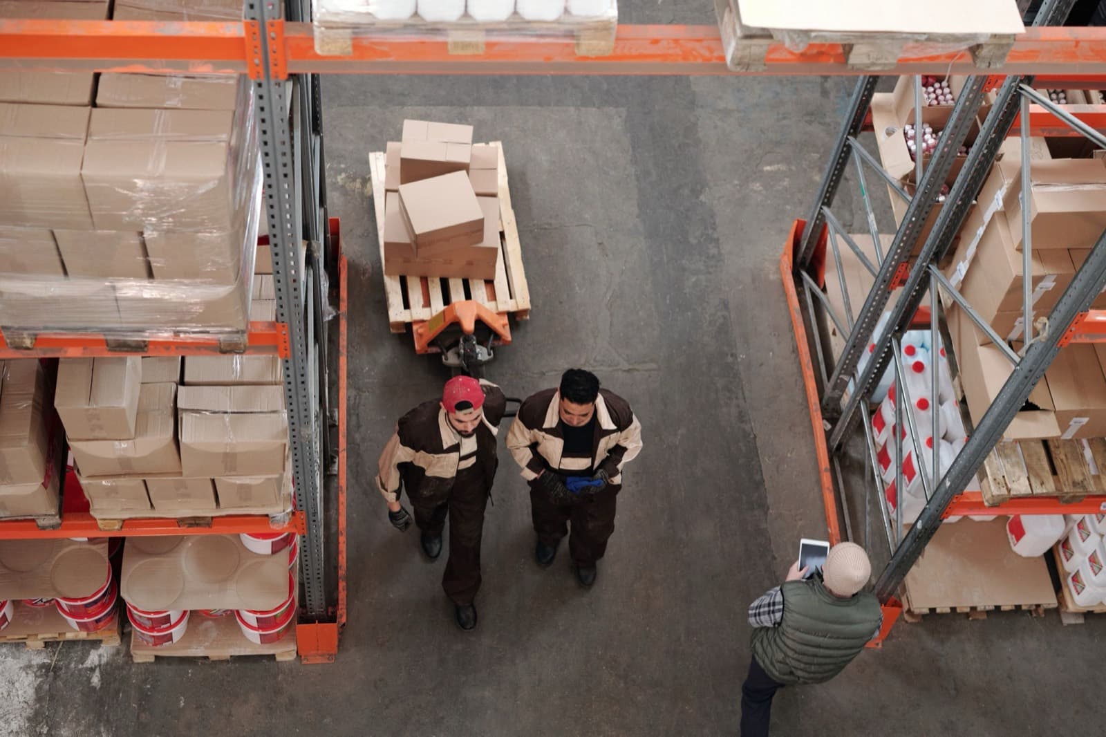 Warehouse workers moving boxed stock on a pallet between racking aisles, seen from above